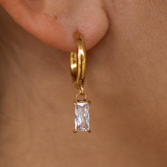 A gold huggie earring with a dangling baguette-cut stone, displayed on a person's ear.