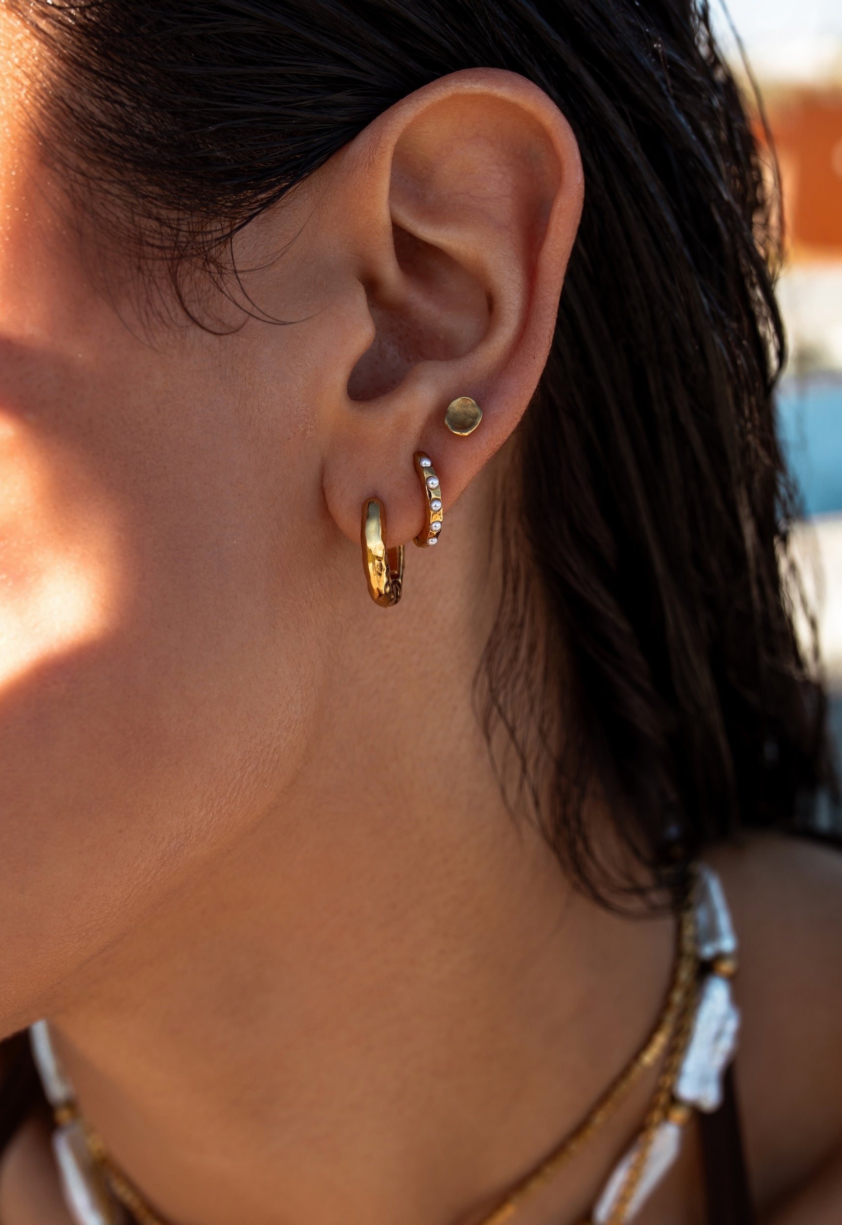 Close up of woman in pool wearing dainty pearl huggies, textured gold hoops, and organic textured gold studs.