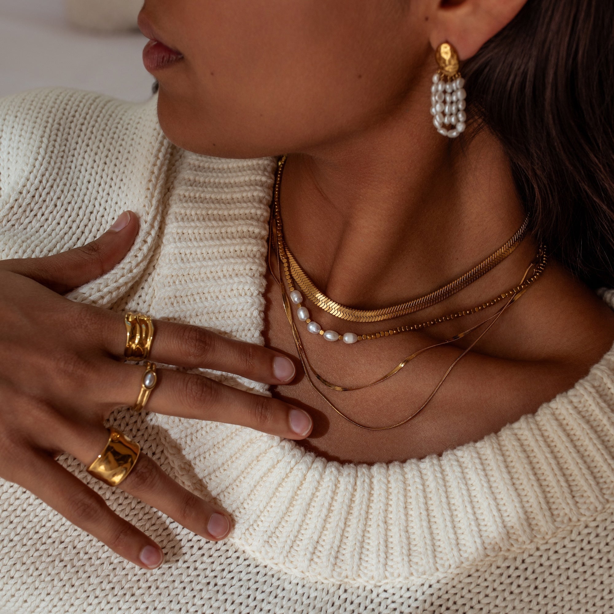 Close-up of a person wearing gold jewelry including necklaces, earrings, and rings on a beige sweater by ALCO Jewelry