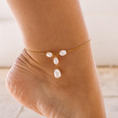 A foot wearing a dainty gold anklet with small white dangling pearls and a gold lobster clasp.
