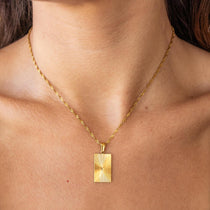 A person wearing a gold-plated necklace with a rectangular sunburst pendant.